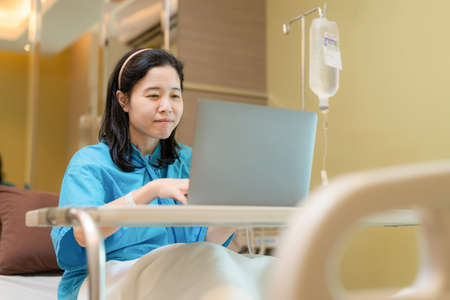 Asia patient woman sitting on the bed with saline solution in hospital ward using laptop, computer...