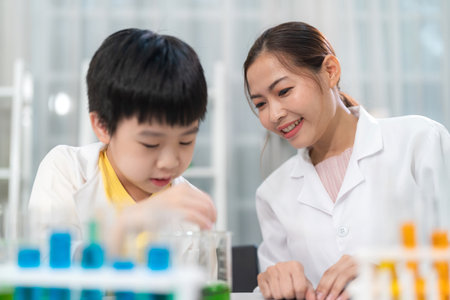 Pretty woman reacher looking at young Asia kid do experiment in laboratory at school. Smart boy use...