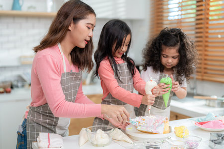 Young little girl standing at counter baking bakery in the modern kitchen at home. Happy smiling parents enjoy weekend. Child and mother wearing apron decorate cream on homemade cake. Learning conceptの写真素材