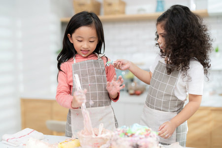 Young little girl standing at counter baking bakery in the modern kitchen at home. Happy smiling and enjoy on weekend. Two kids wearing apron feed cake to each other. Homemade bakery conceptの写真素材