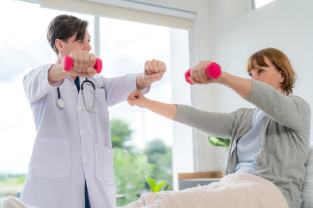 Caucasian elderly patient woman sitting on bed and lifting dumbbell to exercise after treatment in hospital. Workout exercise with doctor. Healthcare medical and physiotherapist Insurance conceptの写真素材
