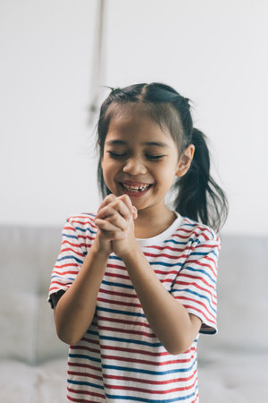 Portrait of happy asian little girl smiling and praying with hands in living room.の写真素材