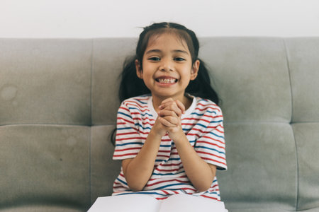 Asian child girl praying with hands  on the white sofa in living room.の写真素材