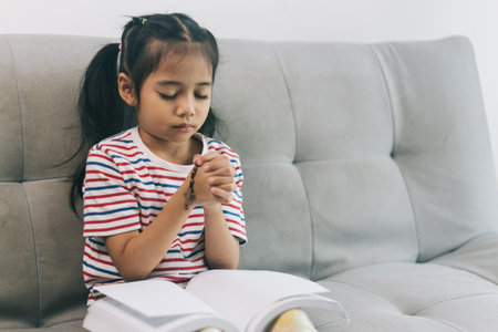 Little asian girl praying for god and reading a book while sitting on sofa at home.の写真素材