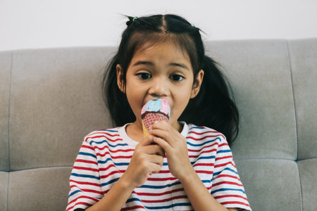 Cute little girl eating ice cream while sitting on sofa at homeの写真素材