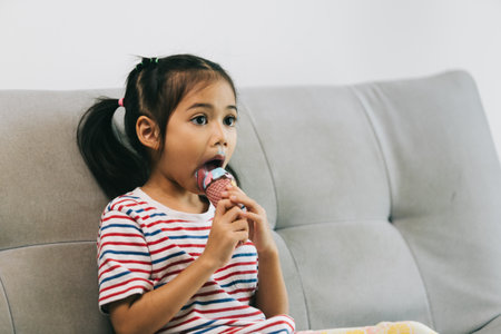 little asian girl eating ice cream on sofa in living room at homeの写真素材