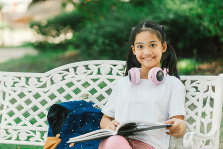 Happy asian little girl reading a book while sitting on a bench in the parkの写真素材