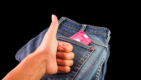 Hand with thumb up before a condom in jeans against black background. Contraceptive concept.の写真素材