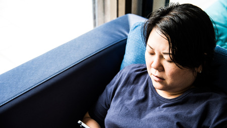 Asian women 40s white skin and plump body in blue dress holding smartphone have a doubt and think gesture on a blue sofa near windowの写真素材