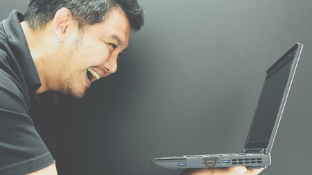 Asian man 40s in black polo shirt holding computer notebook or laptop with laughing and smile gesture in happiness or cheerful concept on black background dark styleの写真素材