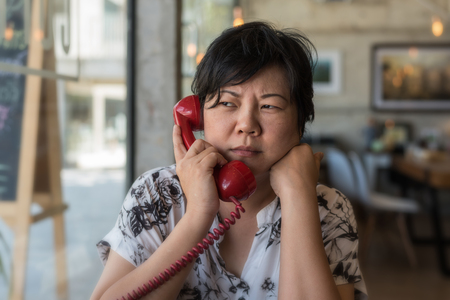 Asian woman 40s white skin in black dress have a doubt and think gesture in a coffee shop cafe with a red telephone vintage styleの写真素材