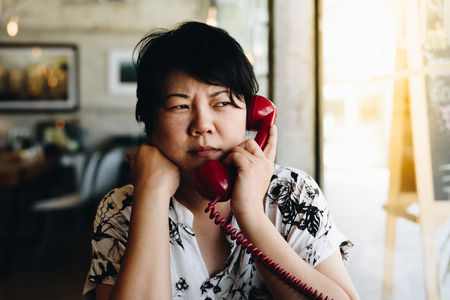 Asian woman 40s white skin in black dress have a doubt and think gesture in a coffee shop cafe with a red telephone vintage styleの写真素材