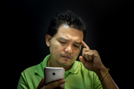 Asian man 40s have a short hair in polo shirt smiling have a thinking gesture with doubt and thoughtful concept using smartphone on black background dark styleの写真素材