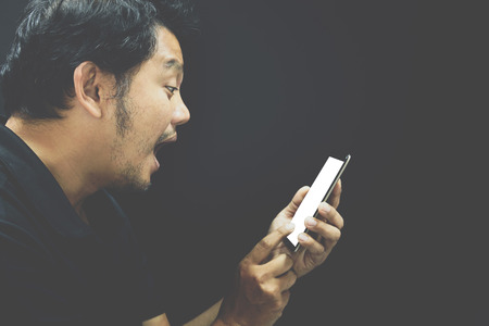 Asian man 40s have a short hair with a beard in black polo shirt stunned and surprise gesture using smartphone on black background dark styleの写真素材