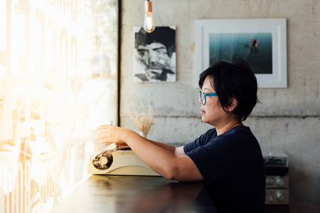 Asian woman 40s white skin with eyeglass sitting for writing something by vintage typewriter in a coffee shop cafeの写真素材