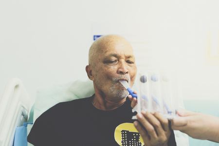 Patient asian elder man 70s use Incentive Spirometer or Triball Tri Flow Meter is medical device to help patients improve the functioning of their lungs on patient bed in ICU. room at hospital.の写真素材