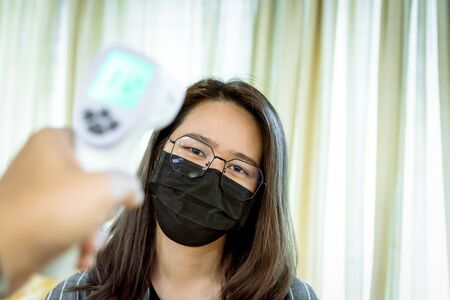 Asian woman wearing mask respiratory protection mask against epidemic flu covid19 or corona virus and body temperature health check measure fever levels by digital infrared thermometer for healthcareの写真素材