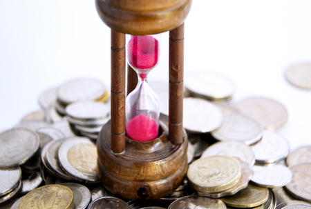 Sand dropping through the bulb of the hourglass and foreign currency coins on background. the...