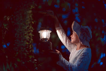 asian girl in a white dress Holding an ancient lantern in her hand, she looked forward in the dark forest at night.soft focus.の写真素材