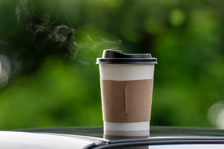 coffee takeaway in a paper cup on top of the car roof green tree background at sunrise in the morning,  selective focus, soft focus.の写真素材