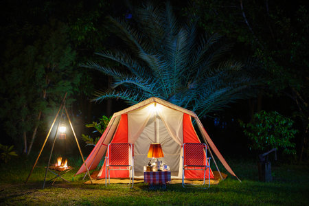 Camping tents at night, vintage cabin tents, antique oil lamps, retro chairs, and a group of camping tents with outdoor coffee-making facilities on wooden tables in a forest camping area.の写真素材
