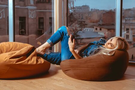 Slim young blonde lying on couch and typing on her mobile phone. Concept of relax, refreshment and leisureの写真素材