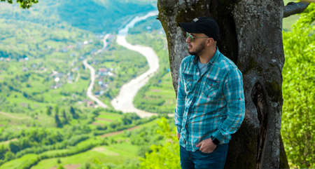 Horizontal of young serious man in sunglasses looking away posing with mountains landscape from...