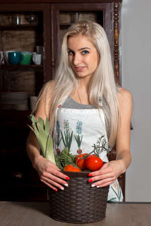 Beautiful girl in an apron holding a basket with vegetables in the kitchenの写真素材