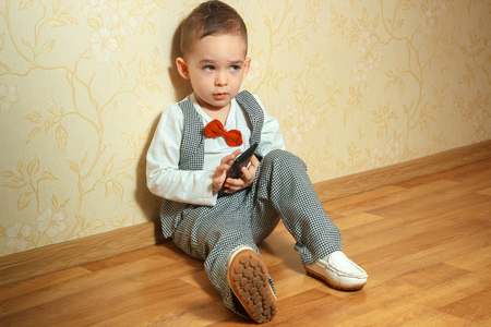boy sits on the floor in elegant suit and red bowtie with mobile phone in his handsの写真素材