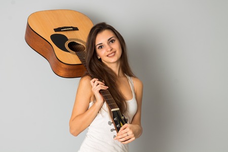 portrait of brunette woman with guitar in hands looking at the camera and smiling in studio on grey...