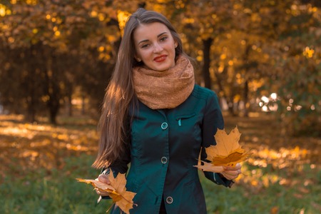 young girl in a warm scarf is standing on the street and keeps the dry leaves in the hands ofの写真素材