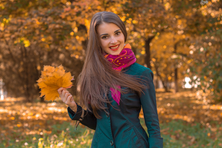 pretty girl in pink scarf around her neck stands in the park looks straight and keeps the leaves in handの写真素材