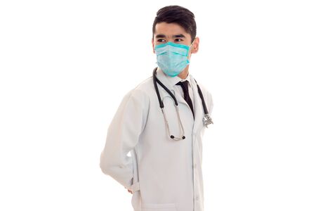 beautiful young brunette male doctor with stethoscope in uniform and mask posing isolated on whiteの写真素材