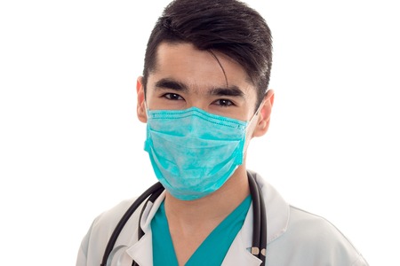 portrait of young brunette man doctor in white uniform and mask with stethoscope looking at the camera isolatedの写真素材