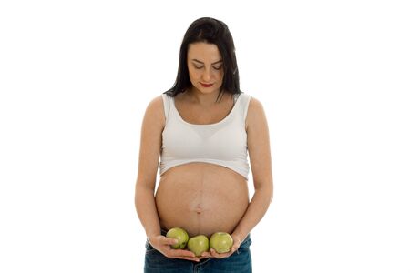 portrait of pregnant brunette in white shirt twith green apples in hands looking down isolated on whiteの写真素材