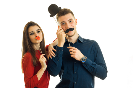 young beautiful girl and Guy hold near face paper props in the form of a mustache, lips and hat and posing for the camera isolated on a white backgroundの写真素材