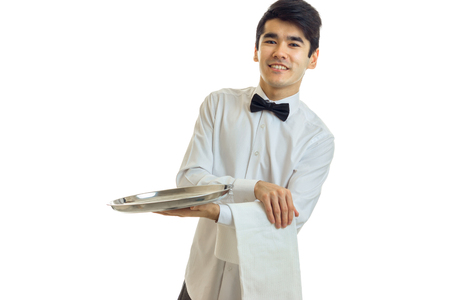 perky young waiter in a white shirt with black bow tie holding a towel and tray for dishware isolated on white backgroundの写真素材