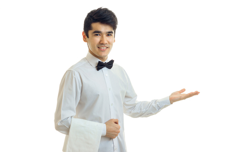 handsome waiter with black hair invites guests to the table with his hand and smiling isolated on white backgroundの写真素材