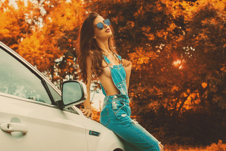 hot brunette in jeans overalls and blue sunglasses sits on a white car in autumn time