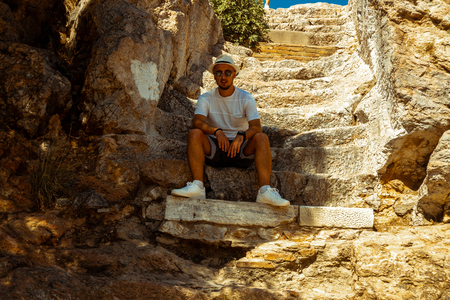 stylish man smiles at the camera sits on ancient greek stairsの写真素材