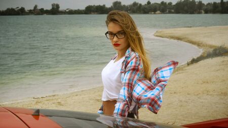 gorgeous blonde in glasses stands at the red car at the seashore