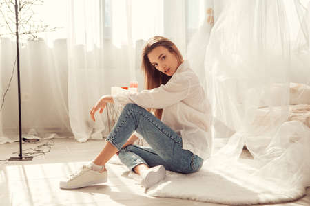 Charming caucasian woman sits in front of bed at light room at homeの写真素材