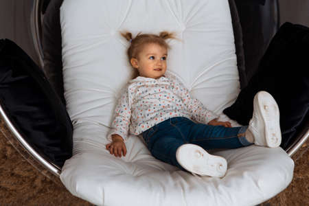 funny little girl sits on white chair and looks aside at homeの写真素材