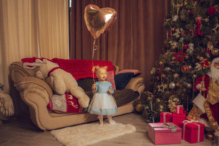 portrait of little baby girl with ballon in her hand looking away with christmas tree behind herの写真素材