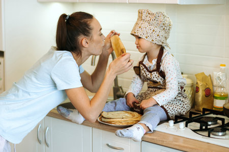 cute little homecook girl with her beautiful mother makes pancakes in white kitchen. Happy family kitchen time.の写真素材