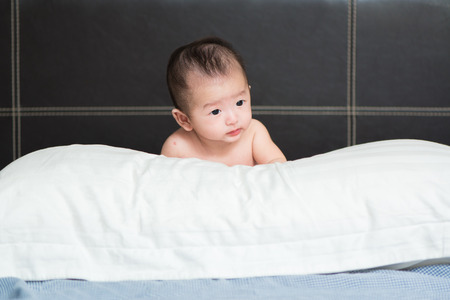 Cute Asian baby about 3 months ages lying, playing, smiling on a white pillow on brown bed and looking to empty spaceの写真素材