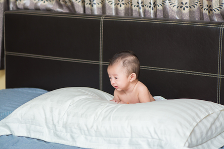 Cute Asian baby about 3 months ages lying, playing, smiling on a white pillow on brown bed and looking to empty spaceの写真素材