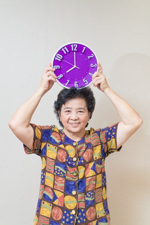Asian senior woman showing and holding purple or violet clock on reddish yellow gray wall background, in studio shot, warm tones with soft shadowの写真素材