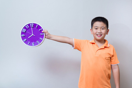 Asian boy showing and holding purple or violet clock in studio shot, on grey wall background with...