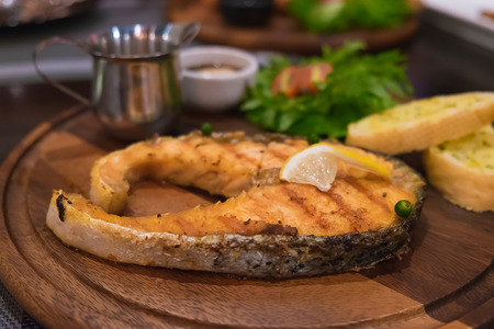 Grilled salmon steak with the fresh salad in white plate. On the wooden table in dark room under incandescent light. shallow focus.の写真素材